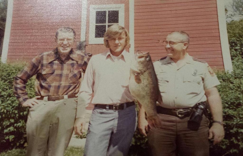Three men standing in front of a house, with the man in the middle holding a very large largemouth bass.