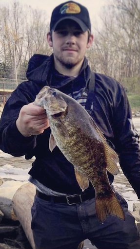 Young man standing on the bank of a river and showing off a 17 inch smallmouth bass that he recently caught.